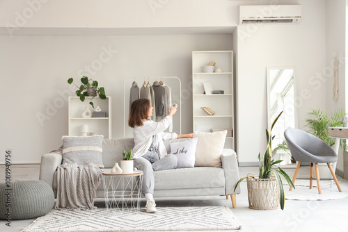 Young pretty woman turning on air conditioner and sitting on sofa in light living room