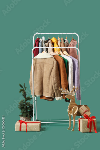 Rack with different clothes, wooden deer, gift boxes and Christmas tree on green background