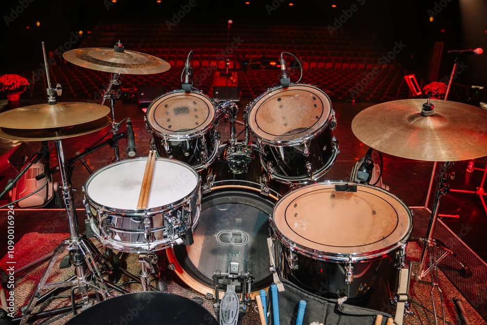 Obraz premium A complete drum set on stage with cymbals, snare, and toms, equipped with microphones, ready for a live music performance in an empty auditorium.