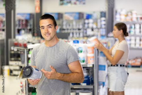 Guy examines can of paint and buys, collects necessary supplies in store according to list for apartment repairs. Blurry female buyer in background photo