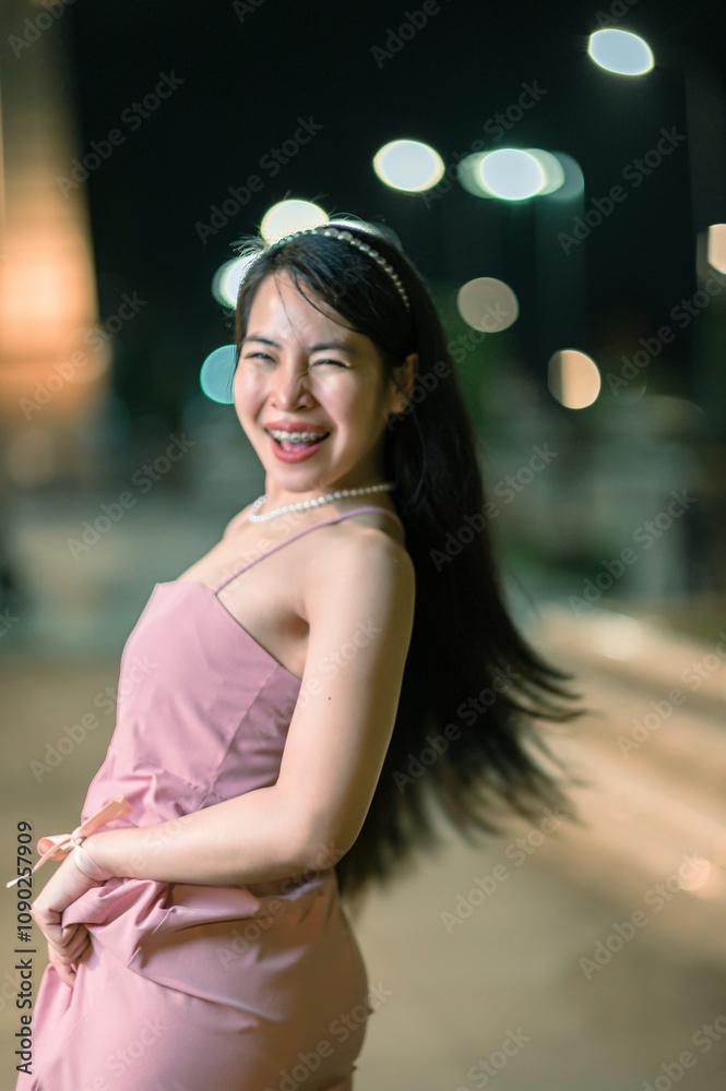 Fototapeta premium Joyful young woman in elegant dress smiles and poses playfully outdoors at night, illuminated by city lights in a vibrant urban setting