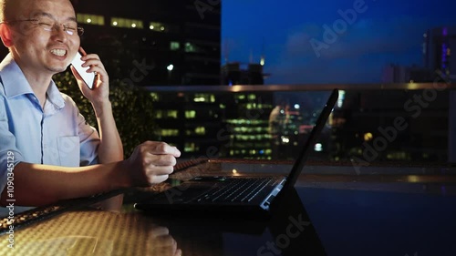 Asian businessman talking on mobile phone with laptop on table on terrace against view of modern city at night