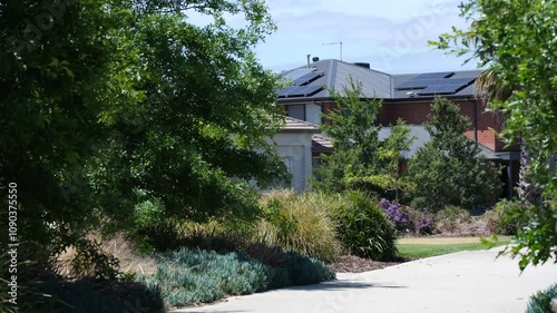 A footpath lined with swaying lush green plants and trees in a public park leads to residential suburban houses in a suburban neighborhood. Wyndham Vale, Melbourne Australia.