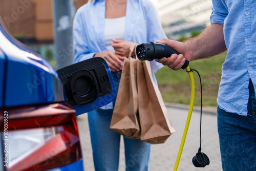 Man plugging in a connector to EV while his wife holds shopping bags at an urban parking lot.