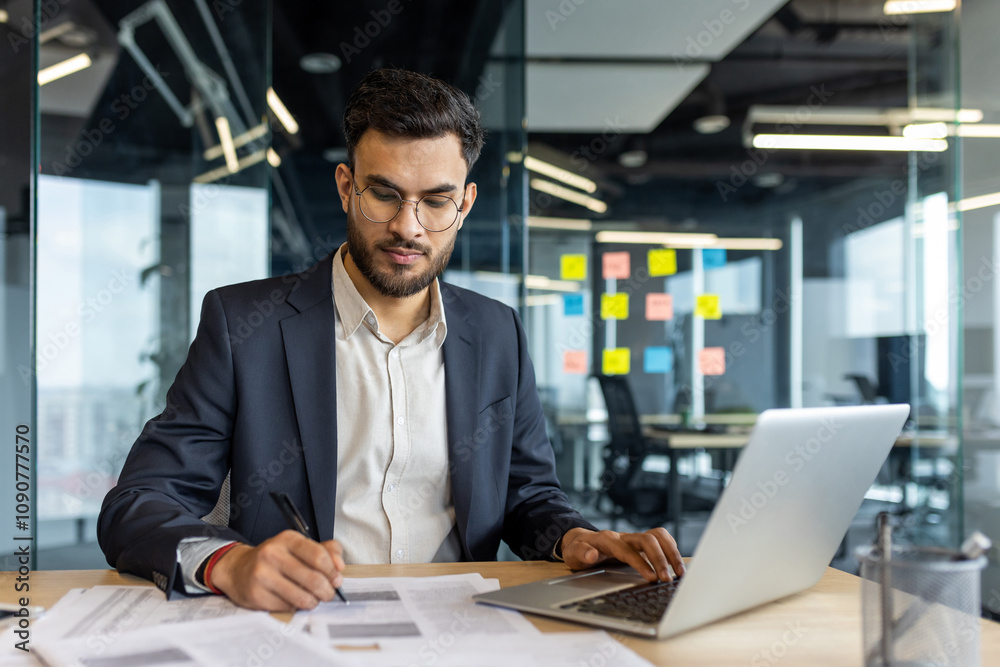 Fototapeta premium Young professional in modern office working with papers at desk, using laptop for multitasking. Businessman wearing glasses focused on tasks, colorful sticky notes adorn glass wall.