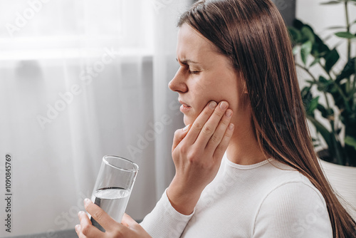 Female sitting on couch at home touching cheek, feel hurt and suffering from sensitive tooth ache. Pain and cavities concept. Close up of young woman feel terrible toothache after drink cold water photo