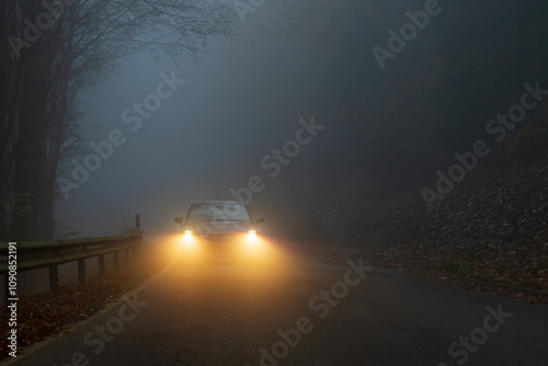 
A car with glowing headlights drives along a foggy forest road at dusk, creating a moody and mysterious atmosphere with low visibility and autumn leaves scattered on the roadside.