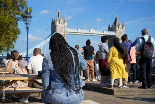 A woman in denim sits on a bench near Tower Bridge in London, surrounded by a diverse crowd enjoying a sunny day. Perfect for concepts of travel, tourism, urban life, and multiculturalism