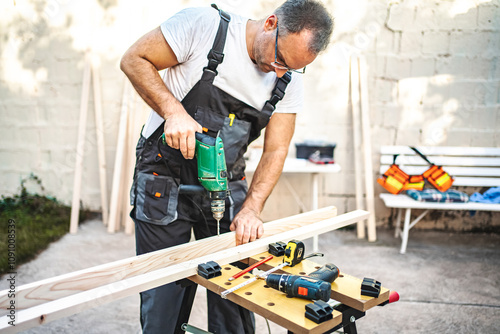 Mature male carpenter using electric drill on a wooden plank outdoors photo