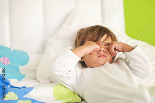 A young boy lies in bed with eyes closed, rubbing his face in a sleepy manner. The bright green walls and colorful bedding create a cheerful morning atmosphere filled with warmth and comfort.