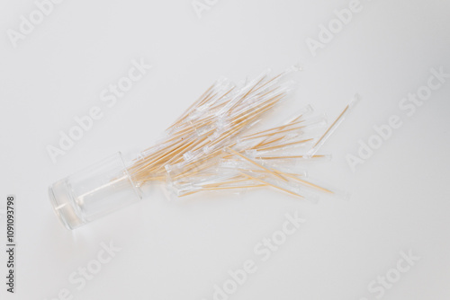 Wooden toothpicks are scattered on a white surface, some falling from a transparent container. The scene is well-lit, highlighting the simplicity of the materials
