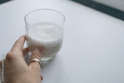A hand adorned with rings gently grips a glass filled with fresh milk, positioned on a bright, minimalist white table in soft afternoon light