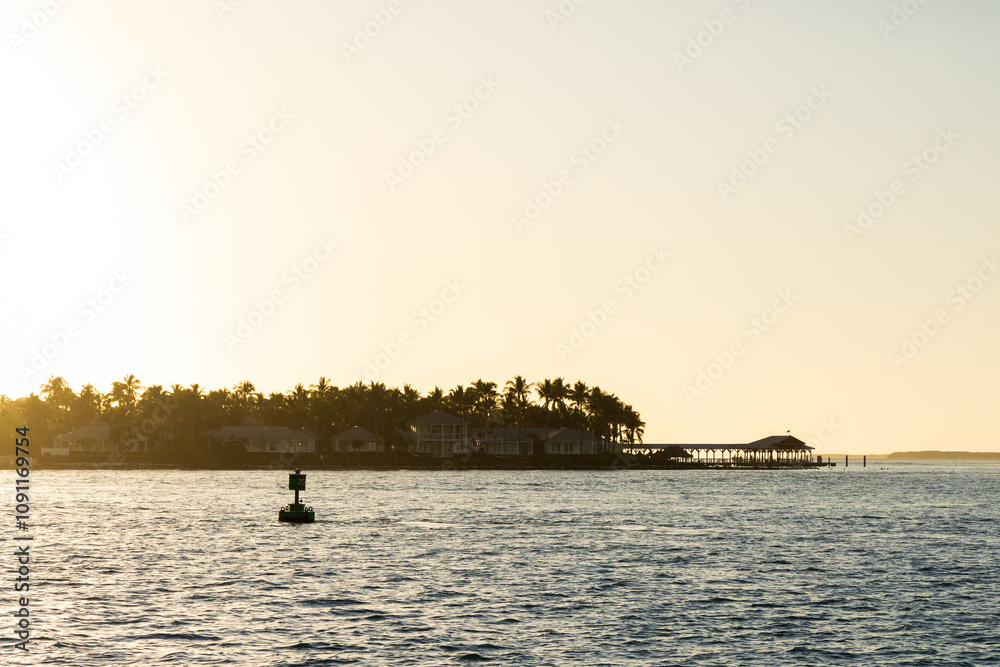 Fototapeta premium A peaceful sunset over the ocean with silhouetted palm trees and a pier extending into the water, bathed in golden light.
