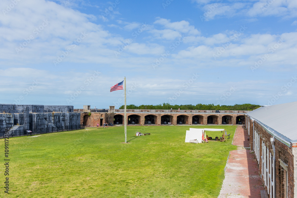 Fort courtyard with an American flag flying, brick archways, cannons, and open grassy space under a bright blue sky, surrounded by historic fort walls. Fort courtyard with an American flag flying, brick archways, cannons, and open grassy space under a bright blue sky, surrounded by historic fort walls.