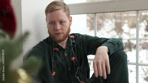 An attractive red-haired, bearded guy is sitting on the windowsill by the Christmas tree.New Year and Christmas concept.