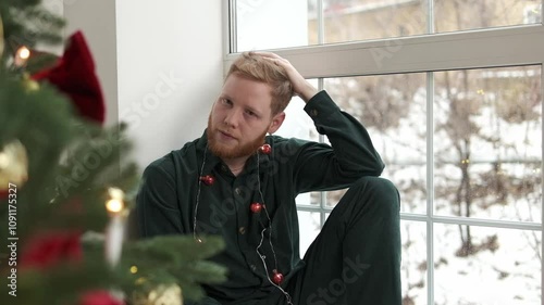 An attractive red-haired, bearded guy is sitting on the windowsill by the Christmas tree.New Year and Christmas concept.