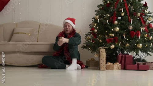 Happy young red-haired man in a Santa hat sitting on the floor next to a Christmas tree.The guy blows up a glitter firecracker.New Year and Christmas concept.