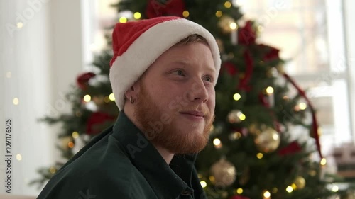 Portrait of a young red-haired man in a Santa hat sitting in front of a Christmas tree. New Year and Christmas concept.