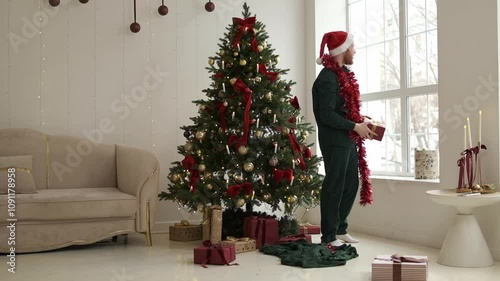 A happy red-bearded young man in a Santa hat unpacking Christmas presents at home by the Christmas tree.New Year and Christmas concept.