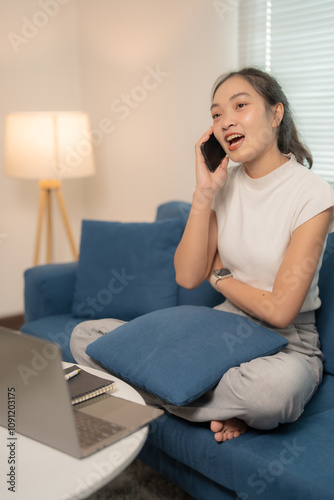 Young asian freelancer is sitting cross-legged on a comfortable sofa, talking on her mobile phone while working remotely from her living room, with a laptop and notebook nearby