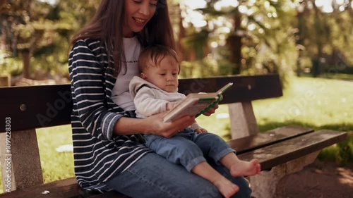 A young mother with a toddler in her arms reads a book while sitting on a park bench during a walk. The concept of upbringing and spending time with a child