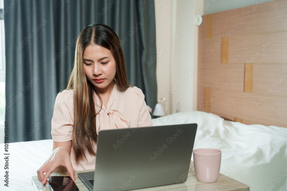 Asian adult woman working at home sitting in cozy bedroom, wearing light pink blouse, holding smartphone and using laptop, warm lighting creating relaxed atmosphere, modern interior design visible. Asian adult woman working at home sitting in cozy bedroom, wearing light pink blouse, holding smartphone and using laptop, warm lighting creating relaxed atmosphere, modern interior design visible.