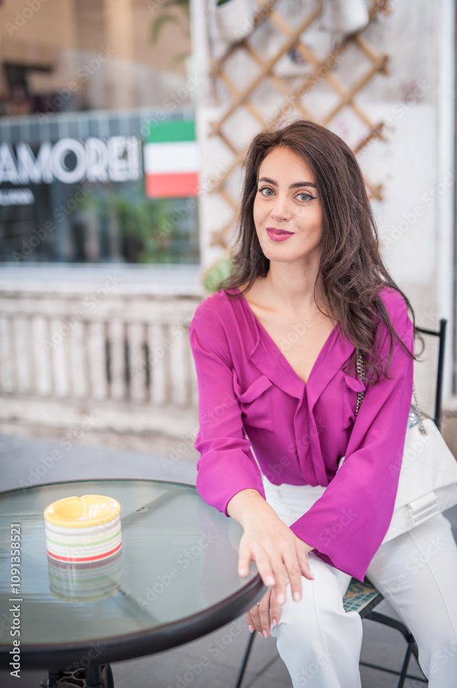 Young beautiful brunette woman in a pink jacket and white pants posing on the street. Portrait of a beautiful young woman in a purple blouse and white pants posing in an outdoor cafe on the street