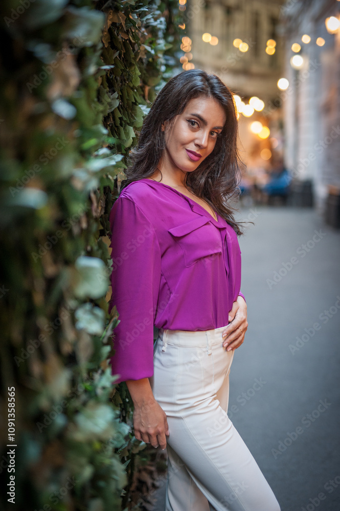Young beautiful brunette woman in a pink jacket and white pants posing on the street. Portrait of a beautiful young woman in a purple blouse and white pants posing in an outdoor cafe on the street