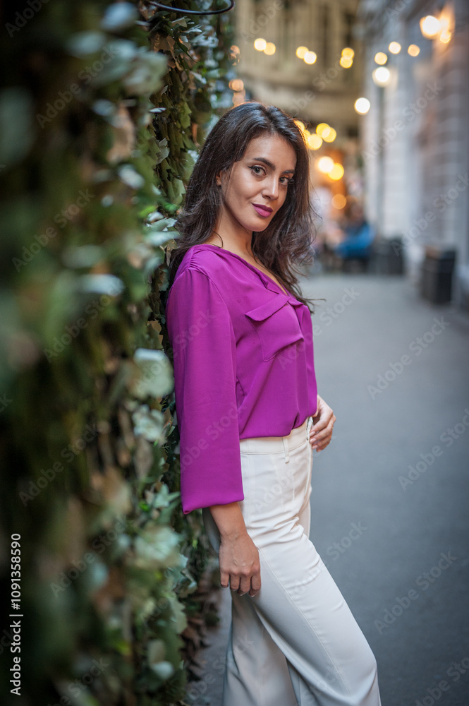 Young beautiful brunette woman in a pink jacket and white pants posing on the street. Portrait of a beautiful young woman in a purple blouse and white pants posing in an outdoor cafe on the street