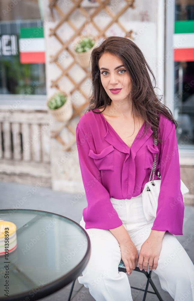 Young beautiful brunette woman in a pink jacket and white pants posing on the street. Portrait of a beautiful young woman in a purple blouse and white pants posing in an outdoor cafe on the street