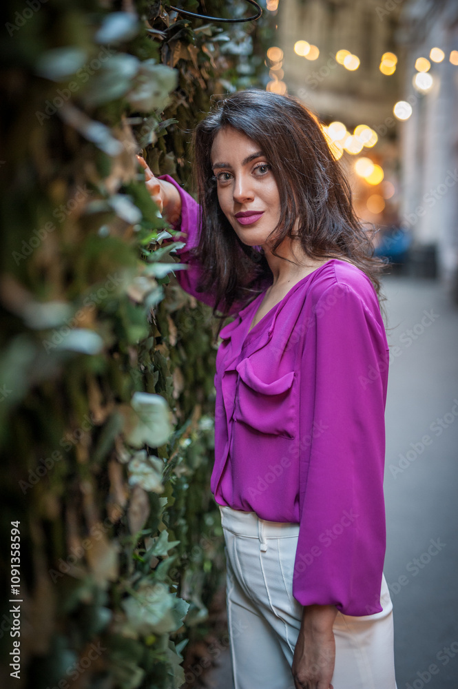 Young beautiful brunette woman in a pink jacket and white pants posing on the street. Portrait of a beautiful young woman in a purple blouse and white pants posing in an outdoor cafe on the street