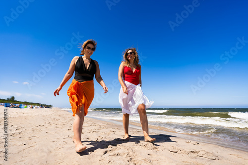 Two beautiful middle aged females wearing one-piece bathing suits and sarongs walking and talking on sandy beach in summertime during holidays. Front view.