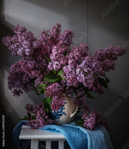 Obraz na płótnie A beautiful arrangement of lilac flowers is displayed in a white vase on a table. The flowers are in full bloom and their vibrant color creates a sense of freshness and vitality. w salonie