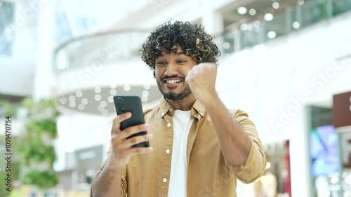 Happy young handsome man rejoices at the good news looking at the smartphone phone standing in shopping center mall store Joyful find sales discount online Surprised male celebrating victory indoor