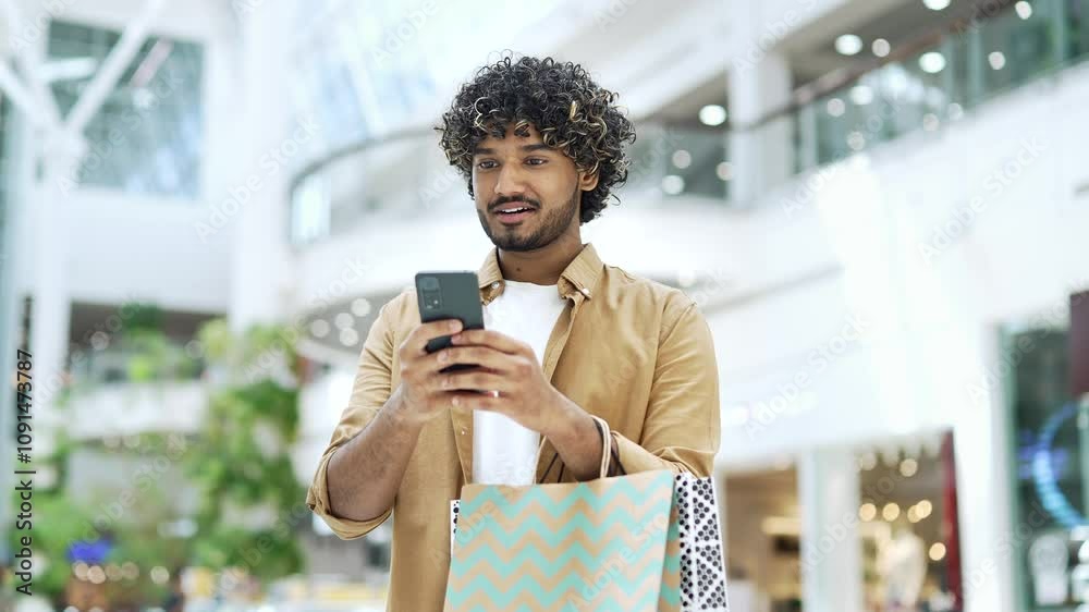Young handsome man happy shopper joyful customer stand in shopping center mall and uses mobile phone browsing chatting typing using smartphone Satisfied glad male tourist in store with paper bag gift