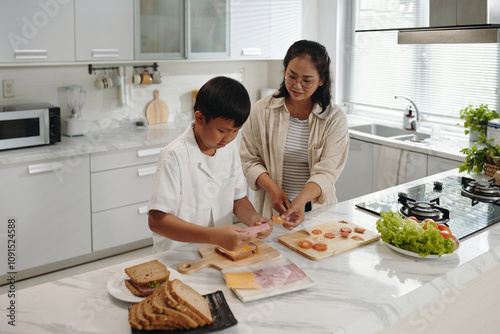 Woman helping young boy in bright kitchen, slicing vegetables and preparing sandwiches on white countertop with fresh ingredients around them. Window letting in natural light photo