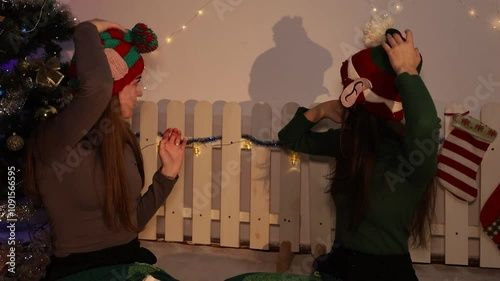 New Year's atmosphere. Two girls try on New Year's hats near the Christmas tree. Subdued cinematic light. Cheerful girlfriends