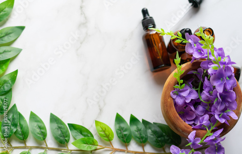 A mortar with some purple flowers and some amber vials of essential oils on a white marble surface. Leaves covering the opposite corners