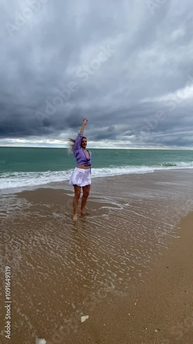 Beautiful blonde woman walking on the beach in autumn. She is wearing a purple shirt and a white skirt, with pearl jewelry on her neck. The woman is running and spinning, enjoying life.