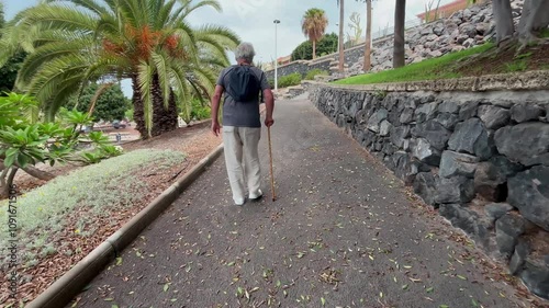 Back view of elder 70s man with walking disability holding knob of cane, leaning on gimp stick walking in the park, suffering from muscles problems, bones disease, recovering after trauma photo