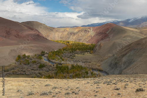 Wide mountain gorge with a stream in the middle. Golden autumn forest grows along the water stream. Mountain slopes are sleep with a red tint like on another planet similar to the Martian landscape.