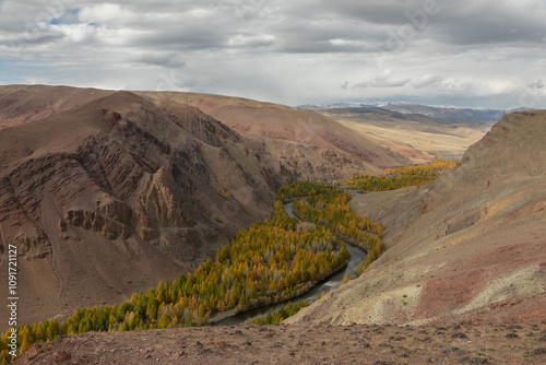 Wide mountain gorge with a stream in the middle. Golden autumn forest grows along the water stream. Mountain slopes are sleep with a red tint like on another planet similar to the Martian landscape.
