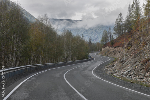 Long curved asphalt road running along the mountain pass. Crash barrier are along the road. Golden autumn roadscape with yellow trees and wild nature. Hilly taiga landscape in the distance