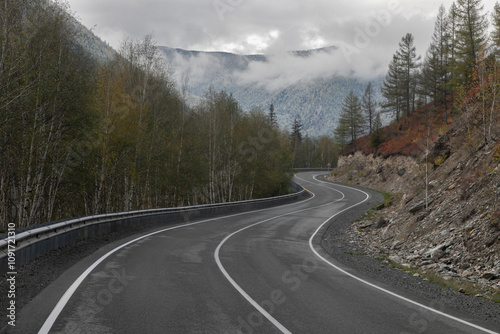 Long curved asphalt road running along the mountain pass. Crash barrier are along the road. Golden autumn roadscape with yellow trees and wild nature. Hilly taiga landscape in the distance