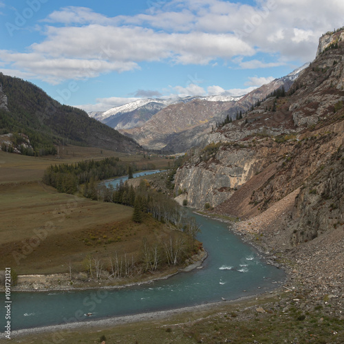 Wide mountain gorge with a beaded turquoise river in the middle. Golden autumn forest grows along the water stream. Rocky steep mountain slopes with a colorful tint