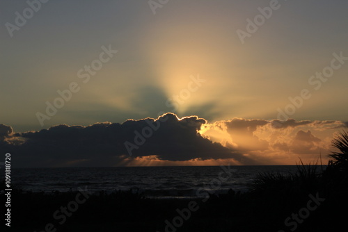 Sunrise over the ocean, sun behind clouds illuminating clouds and outstretched sun rays 