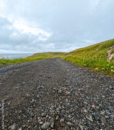 Surface level views of a rocky gravel road or path leading through a grassy Green mountain. The North Atlantic Ocean can be seen in the distance. Cloudy blue sky above. Nólsoy, Faroe Islands. photo