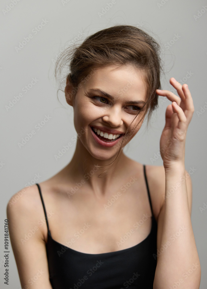 Obraz premium Joyful young woman with tousled hair smiling and touching her face, wearing a simple black tank top against a soft, neutral background. Serenity and beauty concept.