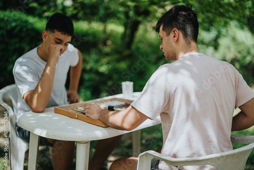 Two young men playing backgammon outside on a sunny day, enjoying a friendly game in the park setting. Relaxing and having fun during a casual outdoor activity.