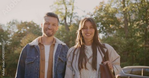 Front view of two people standing together. Looking at each other before glancing at camera. Car parked in front of tall trees in background. Family enjoying fresh air in park. Sunshine.